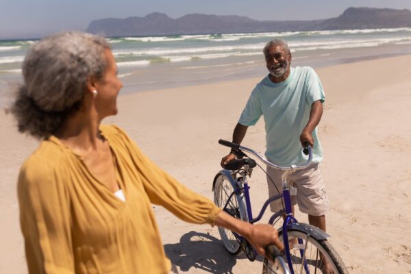 Happy senior couple standing with bicycle and looking each other on beach in the sunshine with mountains in the background