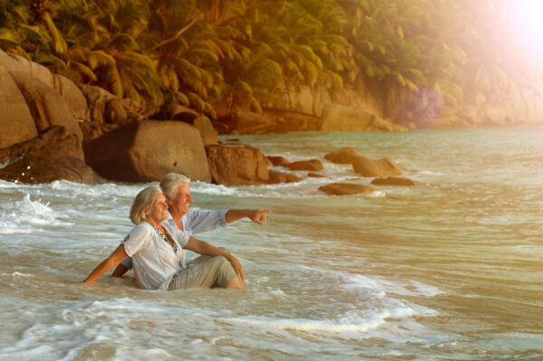 Retired Couple Traveling sitting im water at the beach with rocks behind them